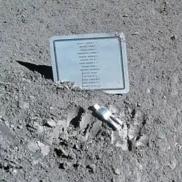 Photographie en couleur d'une petite sculpture argentée accompagnée d'une plaque commémorative placée sur le sol lunaire.