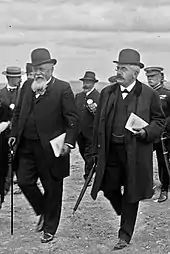 Deux hommes portant chapeau et nœuds papillon, avec chacun des documents sous le bras gauche, marchent côte à côte : l’homme à gauche, à la moustache et à la barbe blanches, s’appuie sur une canne ; l’homme à droite a une moustache noire, porte des lorgnons et tient un parapluie ; à l’arrière-plan, six autres hommes sont distinguables