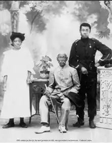 Photo de famille avec une femme en robe blanche à gauche, un soldat africain en uniforme âgé et assis au centre, un jeune soldat médaillé en uniforme à sa droite.