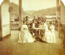 un homme et trois femmes assises autour d'une table avec deux serviteurs noirs debout en arrière-plan