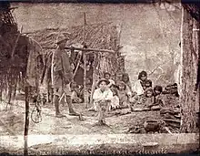 Une vieille photo représentant un groupe de personnes mal vêtues devant une petite hutte construite avec de petites branches