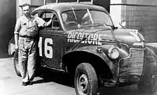 Photo de Juan Manuel Fangio accoudé, debout, à côté d'une vieille voiture.