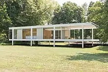 Photographie en couleur d’une maison de campagne moderne entourée de verdure. Maison sur pilotis, structure métallique et façades constituées de baies vitrée.