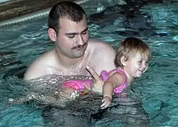  Un homme moustachu dans une piscine tenant dans ses bras une enfant en maillot rose