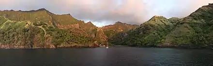 Photographie au soleil couchant de la côte à Hanavave. Il y a quelques bateaux au centre, encadrés de montagnes.