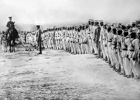 Photographie montrant des soldats mexicains debout et alignés, vêtus d'uniformes beige, passés en revue par un gradé monté à cheval.