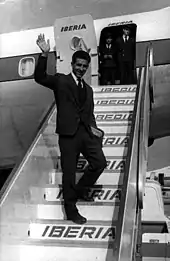 Photographie en noir et blanc d'un homme en costume saluant la foule dans les marches pour accéder à un avion de la compagnie Iberia.