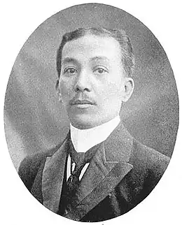 Photo en noir et blanc. Buste d'un homme dans la trentaine, fine moustache, costume et cravate, cheveux courts peignées en arrière