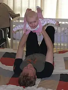 Un jeune père allongé sur le dos sur un édredon sur le sol. Il tient sa fille bébé au-dessus de lui avec ses bras tendus et ses mains autour de sa cage thoracique. Le bébé a ses bras et ses jambes tendus et arque son dos, il sourit à l'appareil photo.