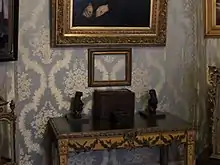 Photo d'intérieur d'un musée, agrandie sur une fine table comportant quelques objets. Au-dessus, un cadre vide laisse apparaître la tapisserie bleue du mur.