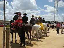 A l'entrée d'une carrière improvisée, un groupe vu de dos de cavaliers en costume traditionnel, certains ayant des jeunes femmes aux robes colorées et froufroutantes en croupe, attendent d'effectuer leur prestation.