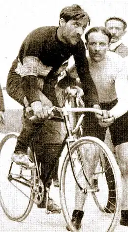 Photographie en noir et blanc d'un homme moustachu, vêtu de noir, assis sur son vélo et entouré de deux autres hommes.
