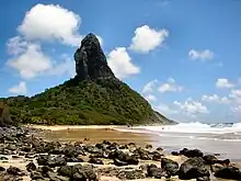 Au premier plan, une mer légèrement agitée à droite et une plage jaune recouverte de pierres noires à gauche. À l'arrière plan, une montagne recouverte d'une végétation verte de laquelle émergent des pans de roche noire.