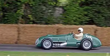 Photographie d'une Formule 1 verte, des années 1950, vue de profil gauche, devant des bottes de paille délimitant un circuit.