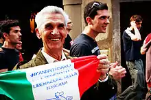 Un homme âgé souriant dans un défilé, tenant un drapeau italien décoré.