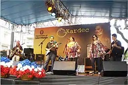 Festival spirite dans les rues de São Paulo en 2010. L'image représente une scène musicale surmonté d'un toit bleu. Des chanteurs et musiciens travaillent devant un panneau géant présentant le portrait d'Allan Kardec et le logo de la municipalité de Sao Paulo.