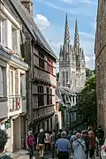 Ruelle bordée de maisons à colombages, et au loin en contrebas, les flèches d'une cathédrale gothique.