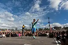 Un homme en T-shirt et short, micro à la main, l'autre bras en l'air, sur une scène musicale, devant une foule.
