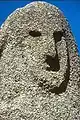 Un visage plat sculpté dans de la pierre grise inhomogène sur fond de ciel bleu.