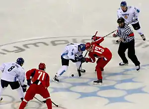 Photographie de cinq hockeyeurs se disputant un palet (trois joueurs en blanc, deux joueurs en rouge) et d'un arbitre en noir et blanc.