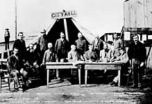 Hommes debout et assis autour de deux tables, faces vers l'appareil. Une grande tente se tient derrière eux avec un panneau de bois où est écrit « City Hall »
