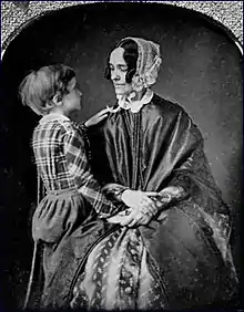 Photographie d'un jeune garçon aux côtés d'une femme assise portant une robe ample