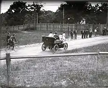 Une voiture sur une piste terreuse. Des coureurs sont visibles derrière elle.