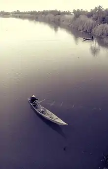 Photo d'un pêcheur dans sa barque vu dans. Il ramène un filet qui était immergé dans l'eau. Plus haut, un autre pêcheur déplace sa barque depuis la berge