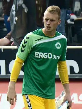 Photographie d’un homme, de pied, en tenue de footballeur, en train de marcher avec un maillot et des gants de gardien de football