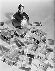 Photographie en noir et blanc d'un flacon de parfum en forme de boule couronnée d'un bouchon, le flacon se trouvant au milieu de petits cubes transparents.