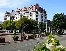 vue en couleurs sur une place publique fleurie avec un bâtiment et une route.