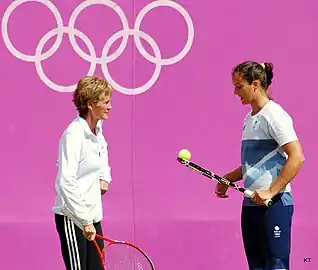 Deux femmes discutent, raquette en main, devant un mur rose marqué du logo olympique.