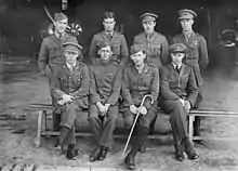 Photographie noir et blanc d'un groupe de 8 hommes en uniforme, formant deux rangs de 4. Le premier rang est assis tandis que le deuxième, derrière, est debout.