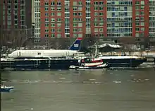 Photo montrant l'avion endommagé, sur la gauche de l'image, après sa récupération du fleuve gelée, au premier plan. L'avion se trouve sur la berge, avec la ville en arrière-plan.