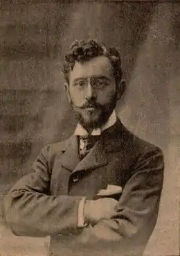 Photographie sépia d'un homme barbu, de face, les bras croisés.