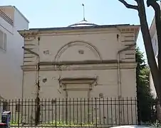 Façade du cabinet d'Histoire Naturelle. Façade sobre dont le crépi blanc sale est fissuré et s'écaille. Des mauvaises herbes tout autour.