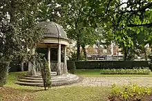 Le Temple de l’Amour vu de gauche, à l’ombre, il est sur la gauche. Sur la droite petite partie du jardin Art déco. En arrière-plan, de grands arbres et des immeubles de l’avenue de Madrid.