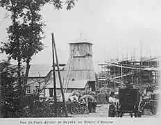 à gauche, un bâtiment en brique et une tour pyramidale en bois sont les installations provisoires. À droite se trouvent des échafaudages autours de bâtiments en construction. Au premier plan se trouvent des chariots et une calèche.