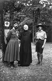 Photo en noir et blanc de trois hommes posant devant quelques arbres : les deux premiers portant une soutane, le troisième est habillé en scout