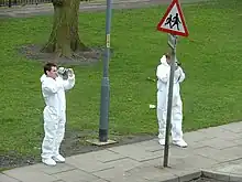 Deux hommes en combinaison blanche, une caméra et un appareil photo à la main tournés vers la droite.