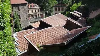 Vue sur un ensemble de toits en tuiles mécaniques constitutifs des bâtiments de la forge Mondière puis au fond 2 grandes bâtisses industrielles
