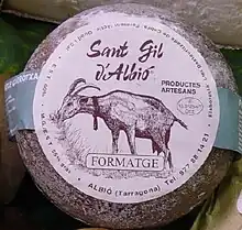 La photographie couleur représente un fromage de chèvre artisanal (indiqué sur l'étiquette) affiné de couleur grise-brun