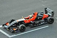 Photographie d'une monoplace de Formule Renault 2.0 noire et orange vue de trois-quarts, d'en haut, sur une grille de départ, dans son ensemble.