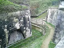 Virage de chemin longeant une barrière, entre deux hauts murs de pierre