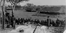 Photo d'un groupe d'Africains assis, devant de grandes cases traditionnelles.