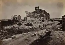 Photographie du fort d'Issy, détruit par la guerre. Il est situé au bord d'une route, sur une butte de gravats entourée d'arbres morts. On ne distingue du fort que des morceaux de façade, aucune d'entre elles n'ayant été épargnée par les bombardements.