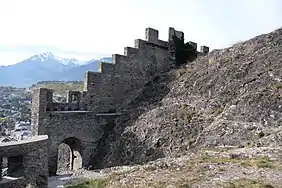 Mur en pierres crénelé suivant le contour d'un rocher muni d'une porte, au sommet de laquelle se trouve un plat accessible depuis la droite.