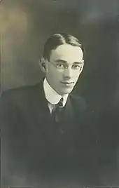 Photographie en noir et blanc d'un homme aux cheveux sombres portant des lunettes et un costume avec cravatte.