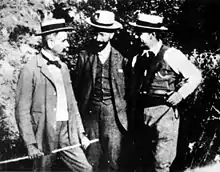 Photographie noir et blanc : trois hommes en chapeau discutent.