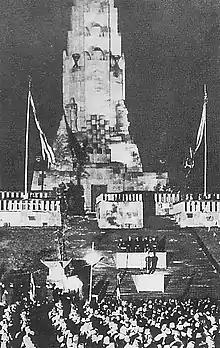 Photo noir et blanc. Cérémonie officielle avec foule devant un escalier menant à un haut monument en forme de tour.